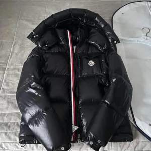 Säljer en snygg och varm svart pufferjacka från Moncler. Jackan har en glansig finish och en cool röd, vit och blå rand längs dragkedjan. Perfekt för vintern med sin quiltade design och långa ärmar. Moncler-loggan syns tydligt på bröstet. Storlek 46