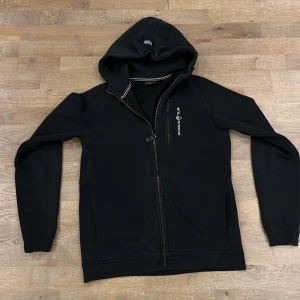 Svart hoodie från Sail Racing - Snygg svart hoodie från Sail Racing med dragkedja och huva. Perfekt för en avslappnad stil. Den har en diskret logga på bröstet och är tillverkad i ett mjukt material för extra komfort. Tröjan är i använd skick