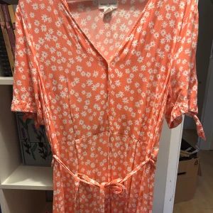 Orange blommig klänning från Holly & White  - Säljer en söt orange klänning från Monki med vita blommor. Klänningen har en v-ringad hals och korta ärmar med knytband. Perfekt för sommarens varma dagar! 🌼