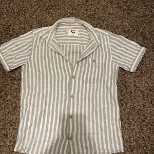 Randig kortärmad skjorta från Cubus - Säljer en stilren randig skjorta från Cubus i storlek 146. Skjortan är kortärmad med knappar framtill och har en bröstficka. Perfekt för en avslappnad look.