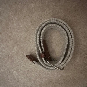 Vit flätat bälte - Snyggt vit flätat bälte med bruna skinnliknande detaljer och en silverfärgad spänne. Perfekt för att ge en stilren touch till din outfit. Passar både till jeans och chinos.