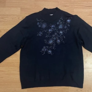 Svart tröja med blommigt mönster från Alfred Dunner - Säljer en svart tröja från Alfred Dunner med ett vackert blommigt mönster i blått och silver på framsidan. Tröjan har långa ärmar och en klassisk passform. Perfekt för kyligare dagar.