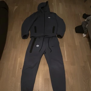 blå mjukisbyxor med huva - Snygga grå mjukisbyxor med matchande hoodie. Byxorna har snörning i midjan och en liten logga på benet. Hoodien har dragkedja och samma logga på bröstet. Perfekt för en avslappnad stil. Storlek M. Hoddie kostar ny 1500 och  byxorna kostar ny 1300 och jag säljer båden hoodien och byxorna tillsammans för 1299