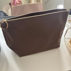 Brun necessär med dragkedja - Snygg brun necessär i syntetmaterial med en praktisk dragkedja och en dekorativ tofs. Perfekt för att organisera dina småsaker. Enkel och stilren design som passar i alla väskor.