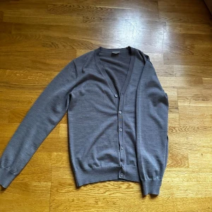 Grå kofta från John Henric - Säljer en stilren grå kofta från John Henric i storlek M. Koftan har en v-ringad design med knappar framtill och långa ärmar. Perfekt för en elegant och avslappnad look.