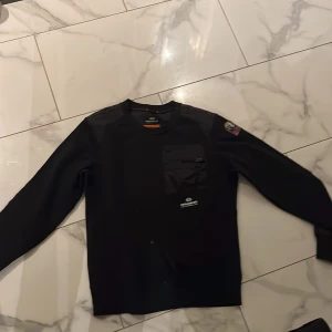 Svart fleece tröja från Parajumpers - Säljer en svart fleece tröja från Parajumpers med en cool ficka på bröstet och ett märke på ärmen. Har en stilren design med långärmade ärmar.
