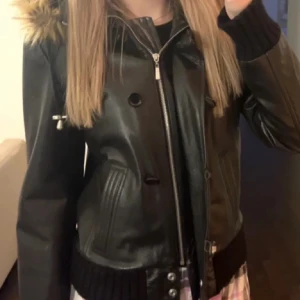 Svart skinnjacka med pälskrage  - Snygg svart skinnjacka i äkta läder med dragkedja och knappar! Jackan har en huva med pälskant och ribbade muddar vid ärmslut och nederkant. kommer tyvärr inte till användning <3