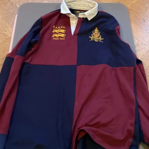 Röd och marinblå rugbytröja från Polo Ralph Lauren - Säljer en klassisk rugbytröja från Polo Ralph Lauren i röd och marinblå färg. Tröjan har broderade emblem och text på bröstet samt en vit krage med knappar. Perfekt för en sportig och stilren look.