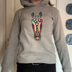 Grå hoodie med färgglatt hästmotiv - Säljer en grå hoodie med ett unikt och färgglatt hästmotiv på framsidan. Tröjan har en klassisk huva och långa ärmar, perfekt för en avslappnad stil. Passar bra till jeans eller leggings.