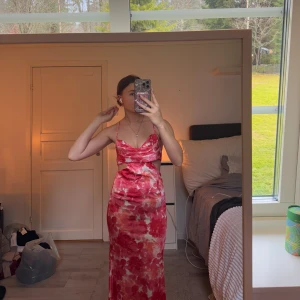 Röd och vit långklänning - Säljer en elegant röd och vit långklänning med blommigt mönster. Klänningen har smala axelband och en öppen rygg med korsade band. Perfekt för sommarens festligheter! Jättefin passform! 