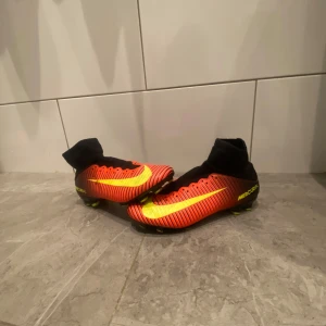Nike Mercurial fotbollsskor - Säljer ett par Nike Mercurial fotbollsskor i röd och gul färg med svart strumpa. Skorna har en modern design med snörning och är perfekta för fotbollsplanen. De har en strömlinjeformad form för optimal prestanda.