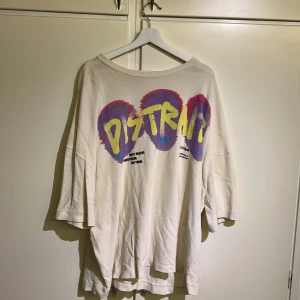 Vit oversized t-shirt från Zara - Säljer en vit oversized t-shirt från Zara med färgglatt tryck i gult, lila och blått. Texten 'DISTRAIT' står i stort på framsidan. Tröjan är storlek XL och perfekt för en avslappnad stil. Köpt för ca 6 månader sedan för ca 400 kr