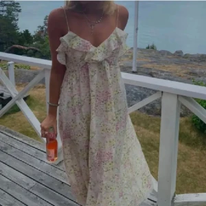 Blommig sommarklänning med volang HM - Charmig blommig klänning med tunna axelband och volangdetaljer vid bysten. Klänningen har en luftig passform och är perfekt för varma sommardagar. Mönstret består av ljusa blommor i rosa, gult och grönt på en vit bakgrund.