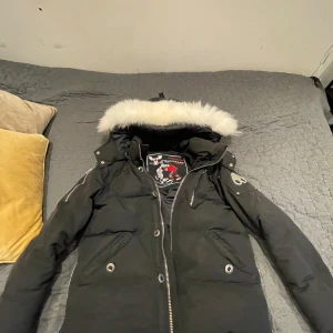 Svart dunjacka från Moose Knuckles - Säljer en svart dunjacka från Moose Knuckles med avtagbar huva och pälsdetaljer. Jackan har dragkedja och knappar framtill samt snygga metalliska detaljer på ärmarna. Perfekt för kalla vinterdagar! ❄️