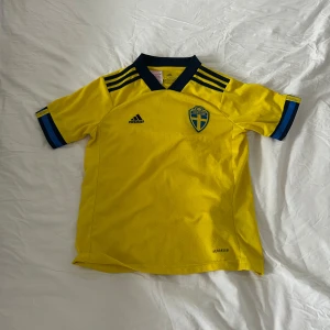 Gul Sverige fotbollströja från Adidas - Säljer en gul Sverige fotbollströja från Adidas med blå detaljer och korta ärmar. Tröjan har Sveriges emblem på bröstet och är tillverkad i AEROREADY-material för bra andningsförmåga. Perfekt för unga fotbollsfans! Kunde inte hitta rätt storlekskategori men det är storlek 11-12 år. Har en liten svart fläck på ryggen