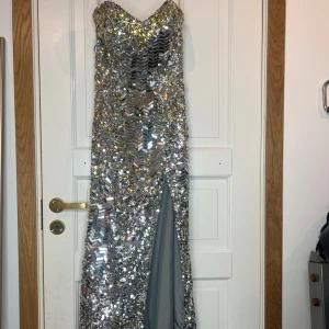 Glittrig silverklänning från Flirt by Maggie Sottero - Säljer en fantastisk glittrig silverklänning från Flirt by Maggie Sottero. Klänningen är axelbandslös med en elegant slits framtill. Perfekt för en glamorös kväll!