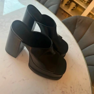 Svarta klackskor med öppen tå - Snygga svarta klackskor med öppen tå och hög platå. Perfekta för att ge lite extra höjd och stil till din outfit. De har en bred klack som ger stabilitet och komfort.oanvända 