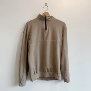 Beige tröja med dragkedja från Santini - Säljer en stilren beige tröja från Santini, tillverkad i 100% bomull. Tröjan har en halv dragkedja framtill och långa ärmar. Perfekt för en avslappnad look.