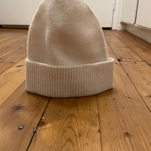 Vit stickad mössa från H&M - Säljer en stilren vit stickad mössa från H&M. Mössan har en ribbad kant och är perfekt för kyliga dagar. Passar de flesta tack vare one size-storleken.