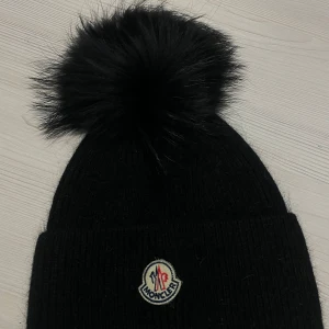 Svart mössa med pälsboll från Moncler - Snygg svart stickad mössa från Moncler med en fluffig pälsboll på toppen. Perfekt för att hålla dig varm och stilren under kalla dagar. Den har Monclers ikoniska logga framtill.