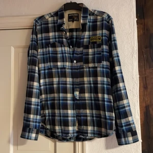 Blå och vit rutig skjorta från Jack & Jones - Snygg rutig skjorta från Jack & Jones i blått och vitt. Skjortan har långa ärmar och knappar framtill. Perfekt för en avslappnad stil med en liten broderad detalj på bröstfickan.