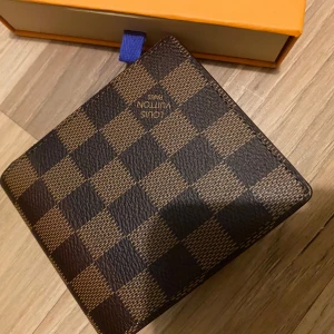 Rutig plånbok - Säljer en stilren plånbok i klassiskt rutigt mönster. Plånboken är brun och tillverkad i skinn, perfekt för att hålla dina kort och kontanter organiserade. Kommer med originalförpackning.