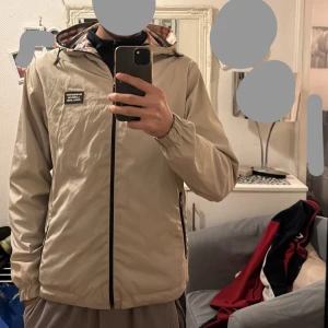 Burberry jacka - Säljer en beige vindjacka från Burberry med avtagbar huva och dragkedja. Jackan är vändbar med ett klassiskt rutigt mönster på ena sidan. Dubbelsidig.