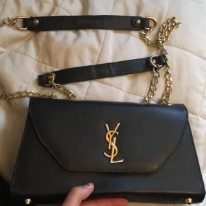 Svart axelväska från Yves Saint Laurent - FÖRSTA PERSONEN SOM SKFIVER TILL MIG EFTER PRISÄNDRINGEN FÅR KÖPA VÄSKAN!! Snygg svart axelväska från Yves Saint Laurent med guldfärgad kedja och logotyp. Väskan har en elegant design med en röd insida och dragkedja. Perfekt för att ge en lyxig touch till din outfit. Ingen aning om den är äkta därför billigt pris