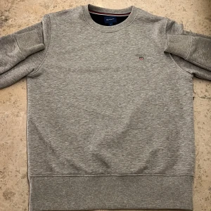 Grå sweatshirt från GANT - Säljer en stilren grå sweatshirt från GANT med rund halsringning och ribbade muddar. Tröjan har en liten broderad logotyp på bröstet. Perfekt för en avslappnad look.