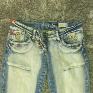 Blå jeans från Crazy Age - Snygga blå jeans från Crazy Age med slitna detaljer och broderade bakfickor. De har en bootcut-stil och är dekorerade med unika mönster. Perfekta för en avslappnad look. De är storlek 36 men är ganska små i storleken. Ett hål på högra knäet och jag är 158 för referens. 