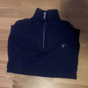 Mörkblå tröja med dragkedja från Gant - Säljer en stilren mörkblå tröja från Gant med halv dragkedja och broderad logga på bröstet. Perfekt för kyligare dagar med sin långa ärmar och bekväma passform.