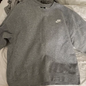 Grå sweatshirt från Nike - Säljer en klassisk grå sweatshirt från Nike med deras ikoniska logga på bröstet. Tröjan har långa ärmar och en rund halsringning, perfekt för en avslappnad stil. Passar bra till både jeans och träningsbyxor.