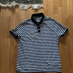 Randig pikétröja från Helly Hansen - Säljer en stilren svartvit randig pikétröja från Helly Hansen. Tröjan har korta ärmar och en klassisk krage med knappar. Perfekt för en avslappnad stil.