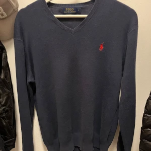 Mörkblå tröja från Ralph Lauren - Säljer en stilren mörkblå tröja från Ralph Lauren i Pima Cotton. Tröjan har en klassisk v-ringning och det ikoniska röda logotypbroderiet på bröstet. Perfekt för en elegant och avslappnad look.