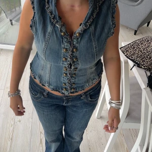 Jeansväst med knappar - Säljer en snygg jeansväst med knappar framtill. Västen har en cool, lite sliten look med fransiga kanter och en tight passform. Supersnygg men behöver rensa💞