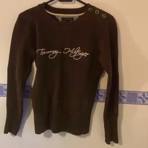 Säljer en snygg brun stickad tröja från Tommy Hilfiger. Den har långa ärmar och en cool detalj med knappar på axeln. Perfekt för höst och vinter, och den är i nyskick. Passar perfekt för en casual look eller när du vill vara lite extra stilren.