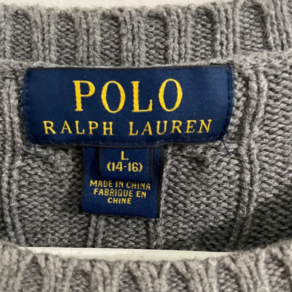 Säljer en snygg grå stickad tröja från Polo Ralph Lauren. Den har ett klassiskt kabelstickat mönster och är perfekt för både höst och vinter. Tröjan är långärmad och har en normal passform. Den är i nyskick och redo att bäras till både vardags och fest!. Tröjor & Koftor.