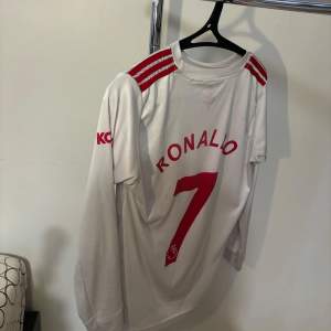 Säljer en vit Manchester United fotbollströja med Ronaldo och nummer 7 på ryggen. Tröjan har röda detaljer och är långärmad. Perfekt för fans av Ronaldo och Manchester United! Har haft den sen 2021 men fortfarande i bra skick! Nypris: 1499kr