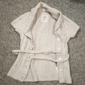 Ser beige ut men är ljus grå stickad kofta från H&M - Säljer en superfin beige eller ljus grå stickad kofta från H&M:s L.O.G.G.-kollektion. Den har korta ärmar och knappar framtill, samt ett bälte i midjan för en snygg passform. Perfekt för vår och höst när det är lite kyligt. Koftan är i bra skick och redo för en ny ägare!💞😊