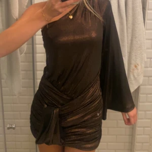 Beun glitteig one-shoulder klänning zara - Snygg brun glittrig klänning med one-shoulder design och knytning i midjan. Perfekt för festliga tillfällen! Den har en asymmetrisk stil med en lång ärm och en kort kjol som ger en elegant look. Endast använd 1 gång. Perfekt till nyår!! 