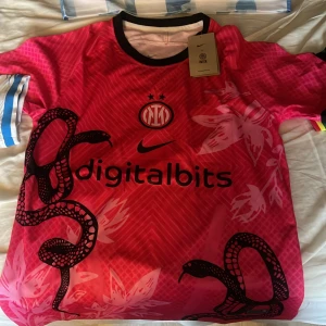 Rosa Inter tröja från Nike - Säljer en cool rosa fotbollströja från Nike med ett unikt ormmönster och blommor. Tröjan har korta ärmar och svart krage. Perfekt för fotbollsfans som vill sticka ut på planen eller läktaren. Märkt med 'digitalbits' och klubbemblem på bröstet.