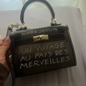 Transparent handväska med text - Säljer en supercool transparent handväska med texten 'UN VOYAGE AU PAYS DES MERVEILLES' och 'SOUVENIR DE L'EXPOSITION 1959'. Väskan har ett guldfärgat lås och en stilren design. Perfekt för att ge din outfit en unik touch!