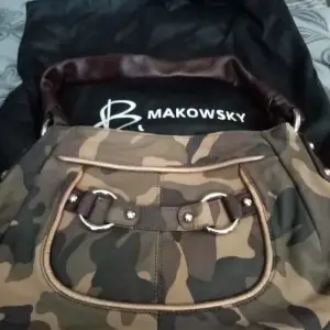 Snygg axelväska i camouflage från B. Makowsky. Väskan har ett coolt mönster och är perfekt för en avslappnad stil. Den har detaljer som dragkedja och metallringar, vilket ger den en tuff look. Perfekt för vardagsbruk eller när du vill sticka ut lite extra.