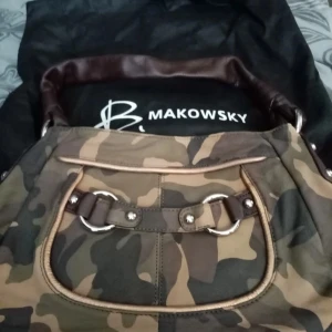 Camouflageväska från B. Makowsky - Snygg axelväska i camouflage från B. Makowsky. Väskan har ett coolt mönster och är perfekt för en avslappnad stil. Den har detaljer som dragkedja och metallringar, vilket ger den en tuff look. Perfekt för vardagsbruk eller när du vill sticka ut lite extra.