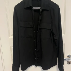 Svart långärmad skjort- jacka - Säljer en stilren svart långärmad skjorta i mycket bra skick. Skjortan har knappar framtill och två bröstfickor med lock. Perfekt för både vardag och fest! 🖤