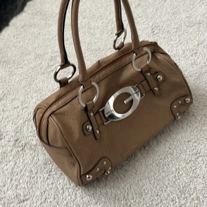 Beige handväska med metallogo - Snygg beige handväska i skinn med en stor silverfärgad metallogo på framsidan. Väskan har dubbla handtag och en dragkedja upptill. Perfekt för både vardag och fest! 