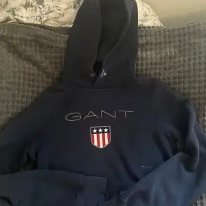 Säljer en snygg mörkblå hoodie från GANT med deras klassiska logga och ett coolt emblem på framsidan. Perfekt för kyliga dagar med sin mjuka och bekväma passform. Den har en stor huva och långa ärmar. Passar både till vardags och chillkvällar. 🧢