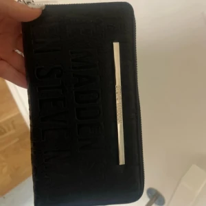 Svart plånbok från Steve Madden - Säljer en snygg svart plånbok från Steve Madden i nytt skick. Den har en stilren design med en dragkedja och en metallplatta med logotypen. Perfekt för att hålla ordning på kort och kontanter. Passar till alla tillfällen!