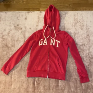 Rosa hoodie från GANT - Säljer en snygg röd hoodie från GANT i bra skick. Den har en dragkedja framtill och en stor luva med vita snören. Perfekt för höst och vår, med långa ärmar och bekväma fickor. GANT-loggan är tryckt i vitt på bröstet. Passar perfekt för en casual look!