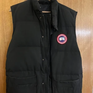 Svart dunväst från Canada Goose - Säljer en svart dunväst från Canada Goose i mycket bra skick. Västen har en dragkedja och knappar framtill samt två stora fickor. Perfekt för höst och vinter när du vill hålla dig varm och stilren. Passar både till vardags och lite mer uppklädda tillfällen. Hör av er om ni har några funderingar eller Liknande🍾